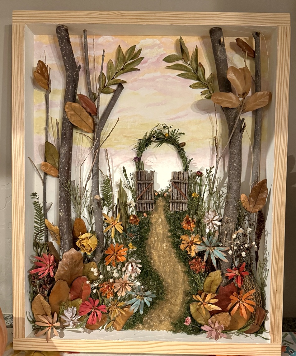 Framed woodland scene with a miniature open garden gate, a winding path, branches and colorful flowers.